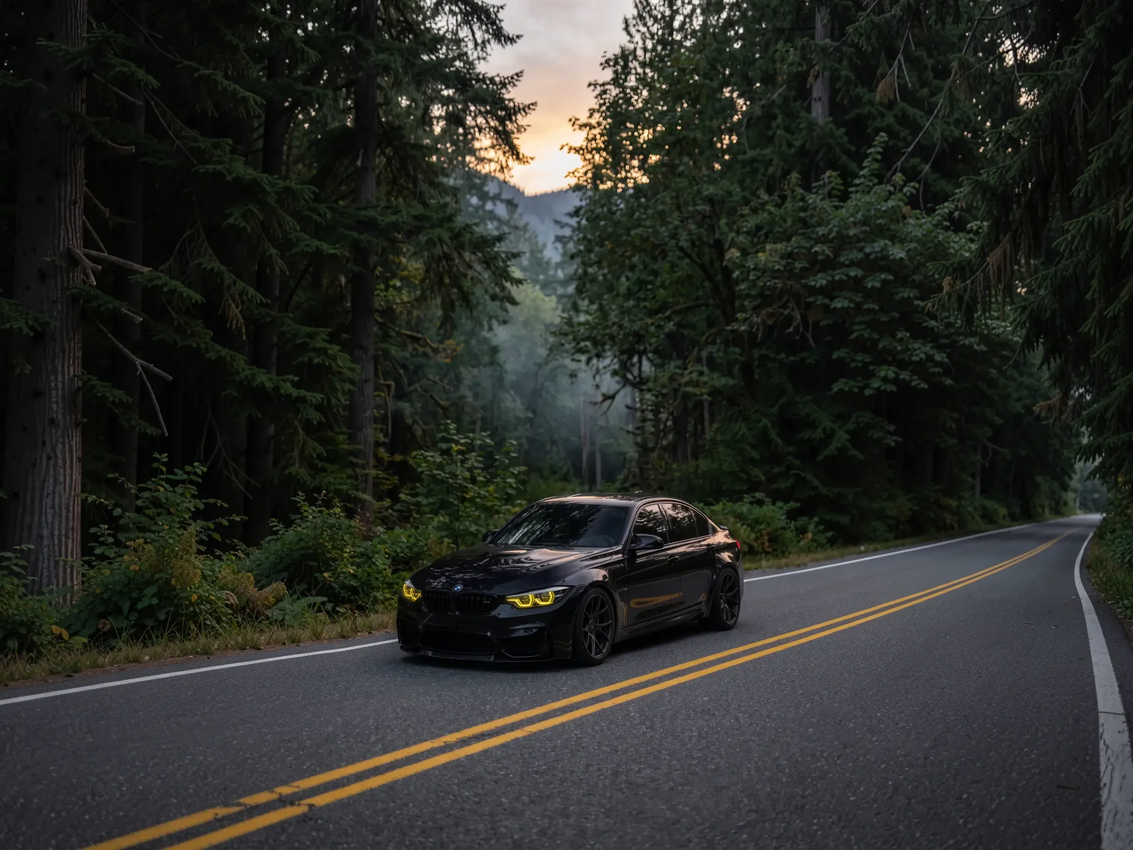 Schwarzer BMW M3 mit eingeschalteten Scheinwerfern auf einer Waldstraße in der Abenddämmerung.